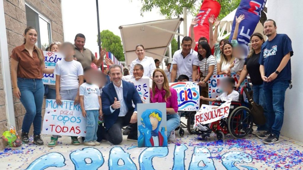 Group of Diverse Adults and Children Posing for a Community Event Photo, Holding Colorful Signs and a Painting, with a Man in a Suit Giving a Thumbs-up. | Tu Colonia Nuevo León