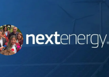 Blue backdrop with the NextEnergy logo; circular inset photo of a woman speaking to reporters at a press conference.