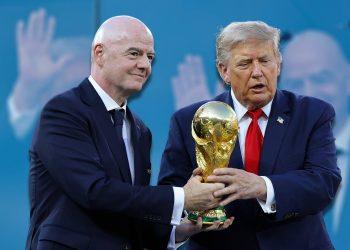 Two men in dark suits exchange a large gold trophy against a blue backdrop, hands clasping the base. This image focuses on the ceremonial trophy transfer.