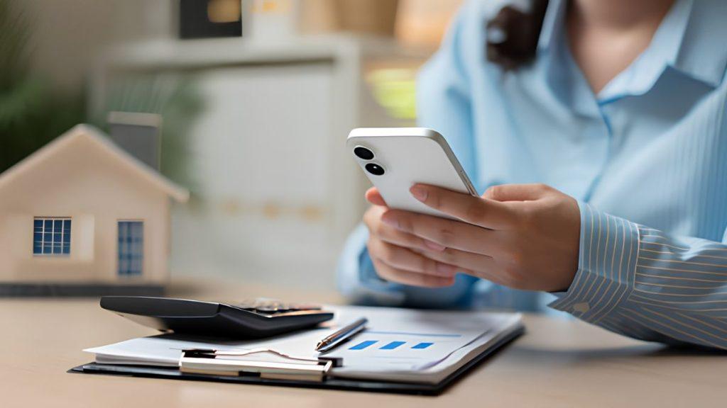 Tips-ahorrar-dinero | Tu Colonia Nuevo León Una Persona Con Camisa De Rayas Azules Sostiene Un Teléfono Inteligente Blanco En Un Escritorio Donde Hay Una Calculadora, Documentos Y Una Maqueta De Una Casa, Lo Que Sugiere Planificación Inmobiliaria O Financiera. | Tu Colonia Nuevo León